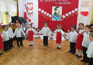 Dziewczynki w stojach biało-czerwonych oraz chłopcy ubrani na galowo w półkolu śpiewają piosenkę patriotyczną.